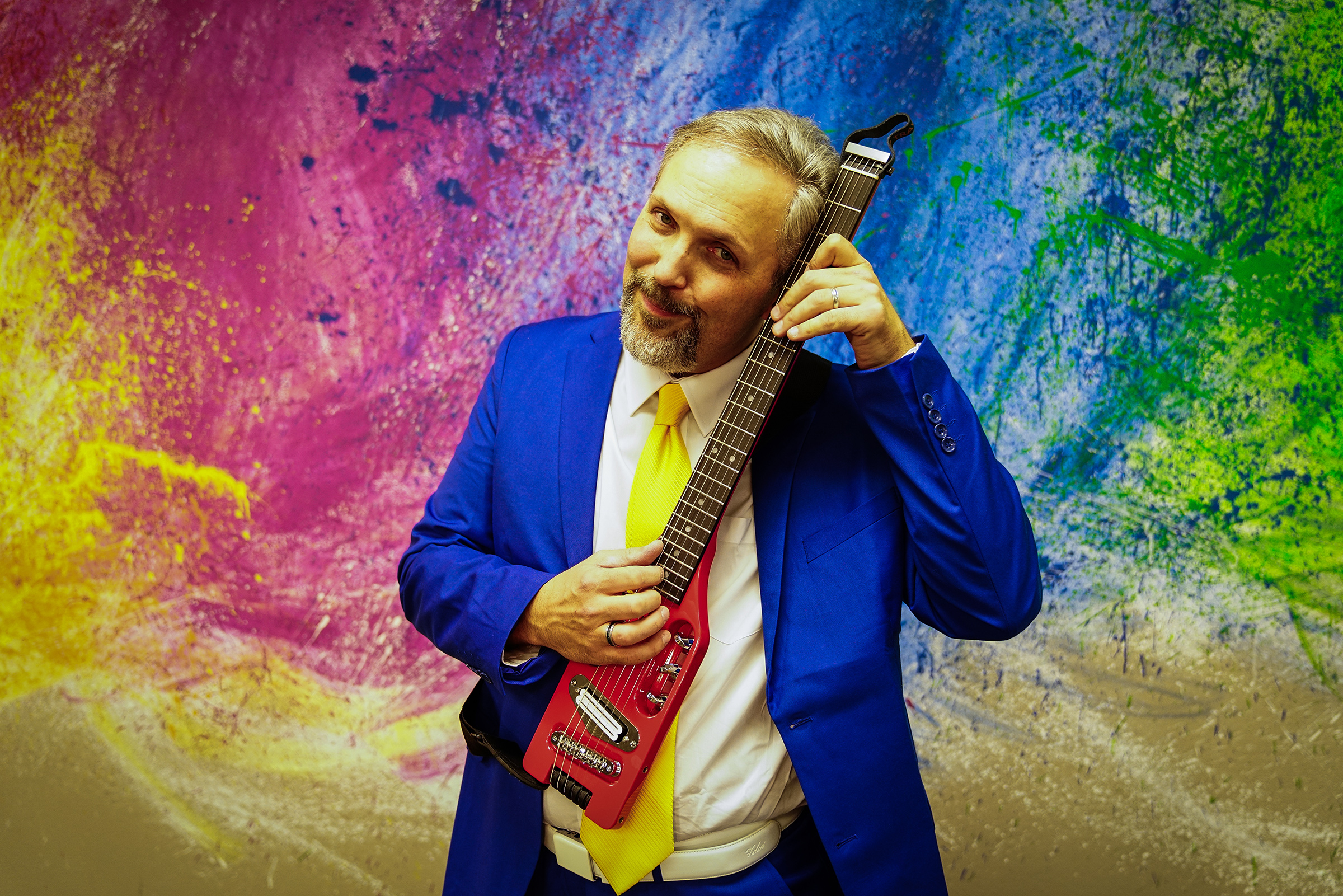 Musician Eric Herman holds a small electric guitar against a colorful abstract background