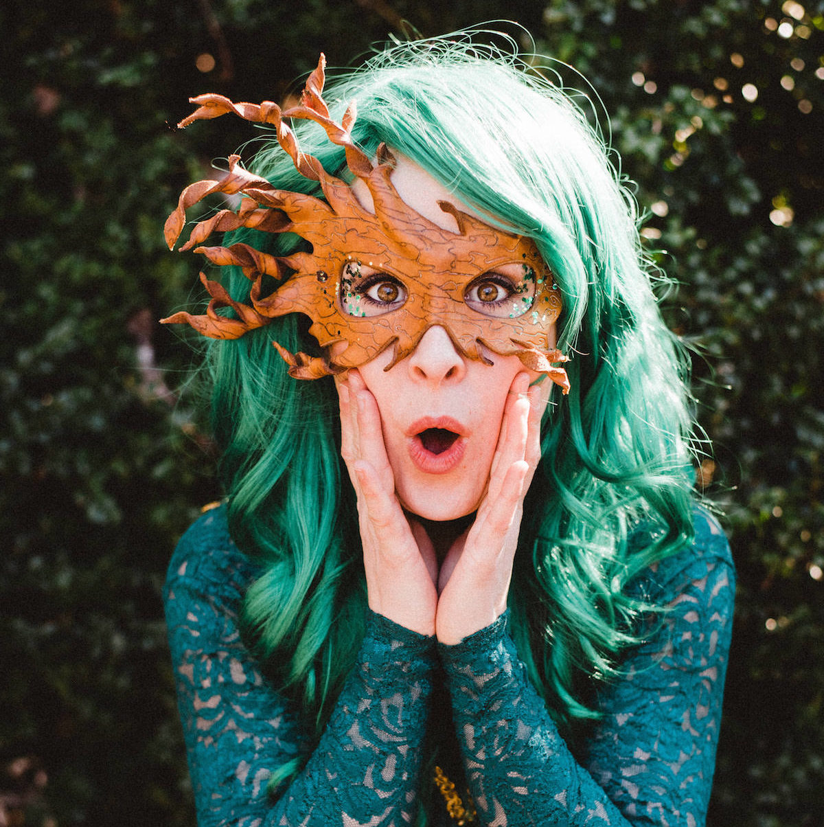 Jessa Campbell with green hair, a nature-themed mask over the top part of her face, eyes and mouth wide open and hands pressed to the sides of her face