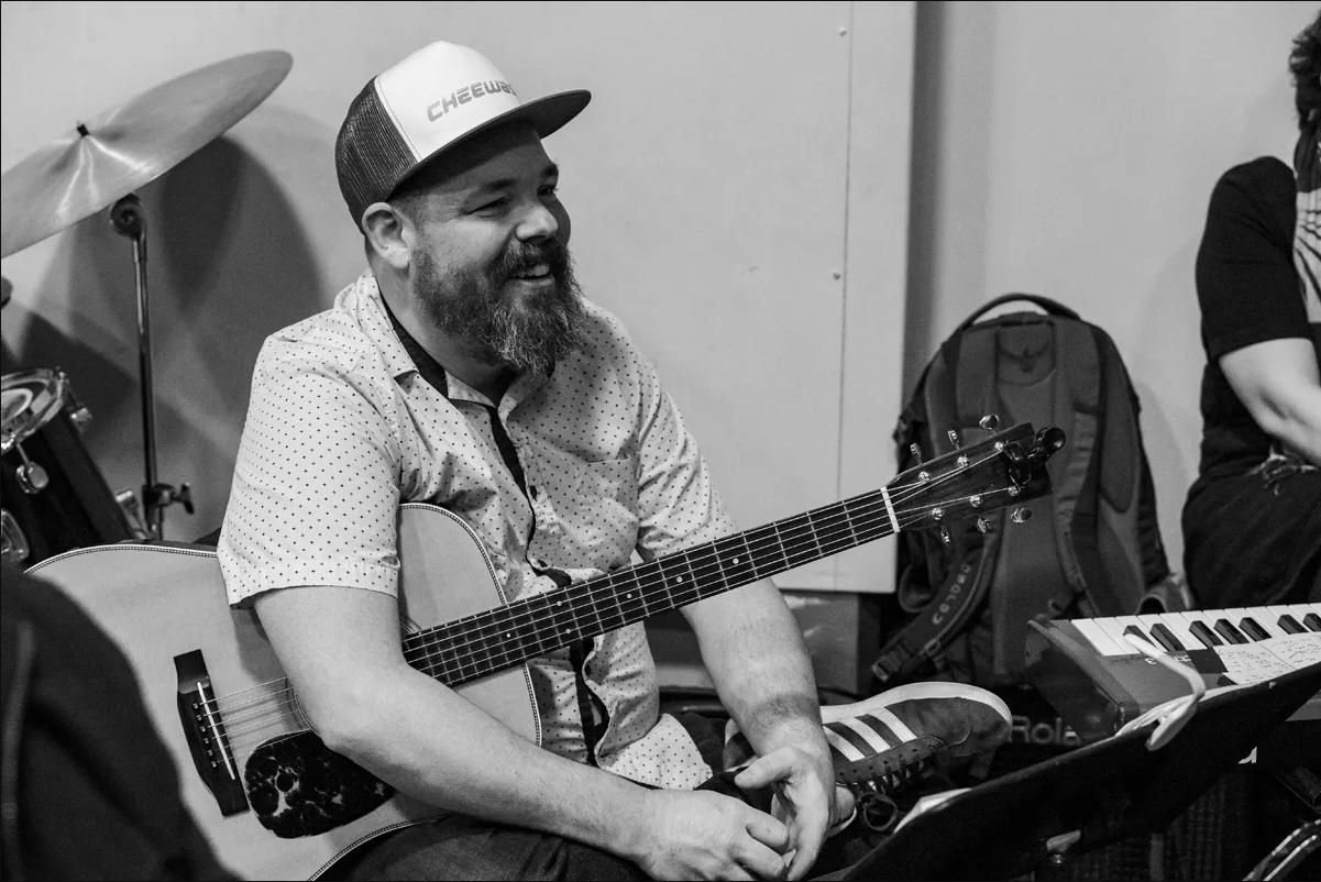 Bearded, smiling man wearing a trucker cap and cradling a guitar