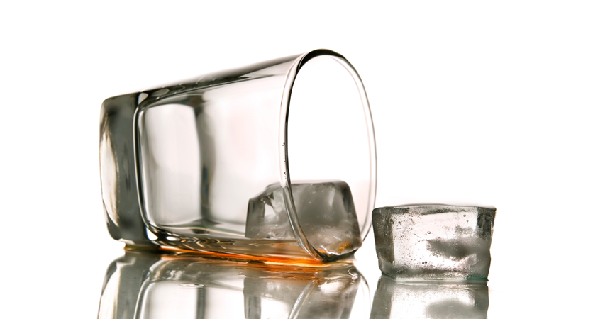 A photograph of a rocks glass lying on its side with two ice cubes tipped out of it. A small amount of brown liquor remains in the glass.