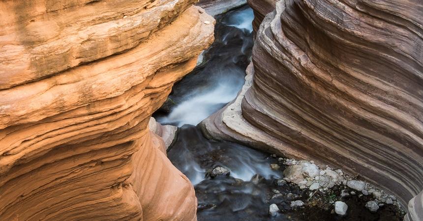 A photo of a space between two rock faces at Grand Canyon National Park. Each rock face has smooth, horizontal grooves etched into them by the Colorado River.