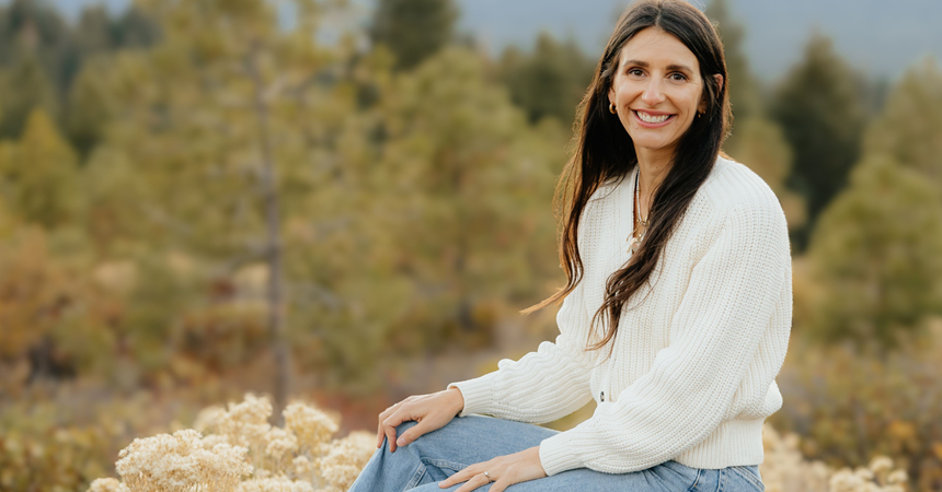 Whitney Wedam, a smiling woman with light skin and brown hair poses on a rock in front of a sagebrush landscape.