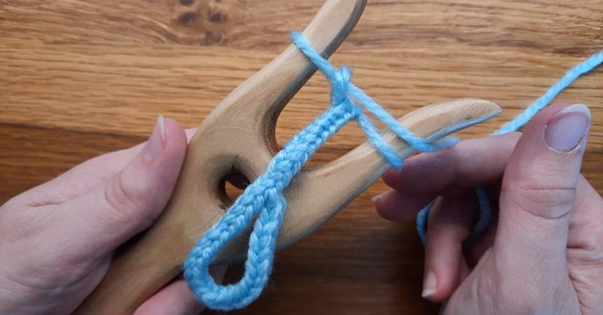 A craftperson's hands braids a cord by winding it on a two-pronged wooden tool.
