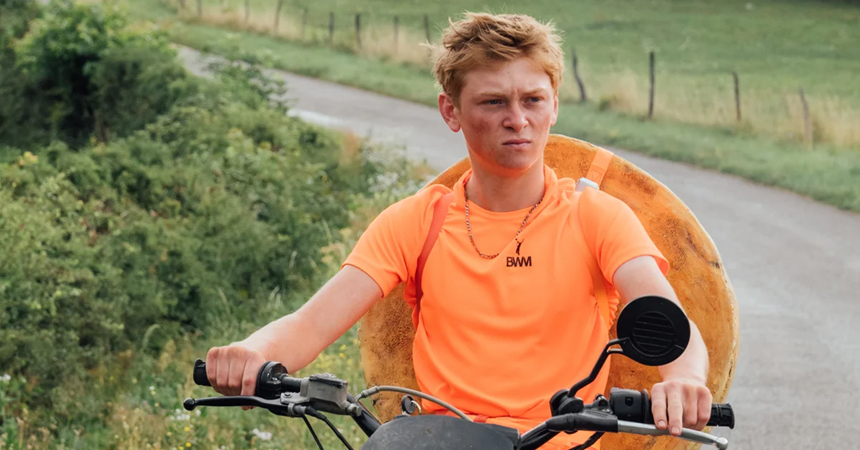A young man rides a motorbike with a large wheel of cheese strapped to his back.