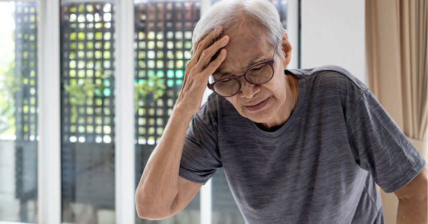 An elderly Asian man stoops with a hand to his forehead, wincing in pain.