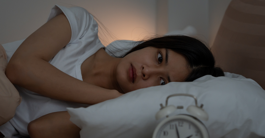 A woman lies awake in bed with an unhappy expression, staring at her alarm clock.