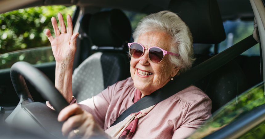 A white woman in sunglasses smiles and waves from the driver's seat of a car.