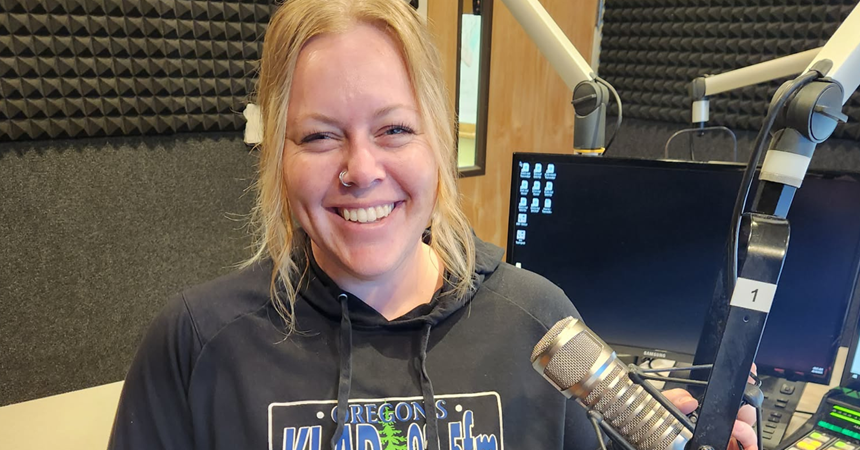 Melissa Tolbert, a light-skinned woman with long blond hair and wearing a KLAD station hoodie, smiles from inside a recording booth.
