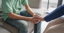 Two people sit across from one another. One person has their hands clasped in front of them while the other lays their hand on the first person in comfort.
