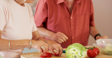 An older man and woman chat while cutting up lettuce and tomatoes for a salad.