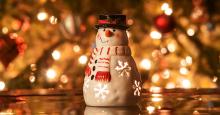 A snowman figurine in front of a blurry background of Christmas lights.