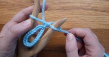 A pair of hands braid a length of light blue yarn with a lucet, a wooden tool with two prongs that hold loops of yarn as it is braided.