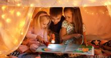 Three children read a picture book with flashlights inside a blanket fort.