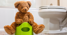 A photograph of a stuffed teddy bear sitting atop a bright green training potty toilet, with a full-size toilet in the background.