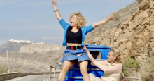 In a scene from Two Tickets to Greece, Magalie (played by Laure Calamy) stands in the back of the open flatbed of a truck cheering exuberantly while Blandine (played by Olivia Cote) tries to get her to sit down.