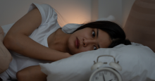 A woman lies awake in bed with an unhappy expression, staring at her alarm clock.