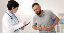 A man doubles over in pain clutching his abdomen while a doctor takes notes on a clipboard.