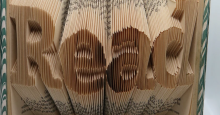 An open book whose pages have been folded so that the edges make a 3d sculpture of the word "read."