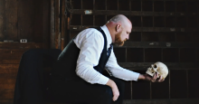 Seth Howard, a bald light-skinned man with a red beard wearing a formal vest and shirt, gazes into the eyes of a replica skull.