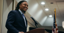 Malcolm Kenyatta, a young Black man with locs and wearing a blue suit, smiles out at the audience from an event podium.