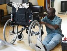 A dark-skinned man sits next to a wheelchair, examining a mechanism within.