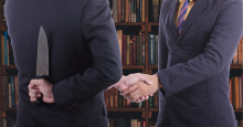 Two men in suits shake hands. One of them is holding a large chef's knife behind his back.